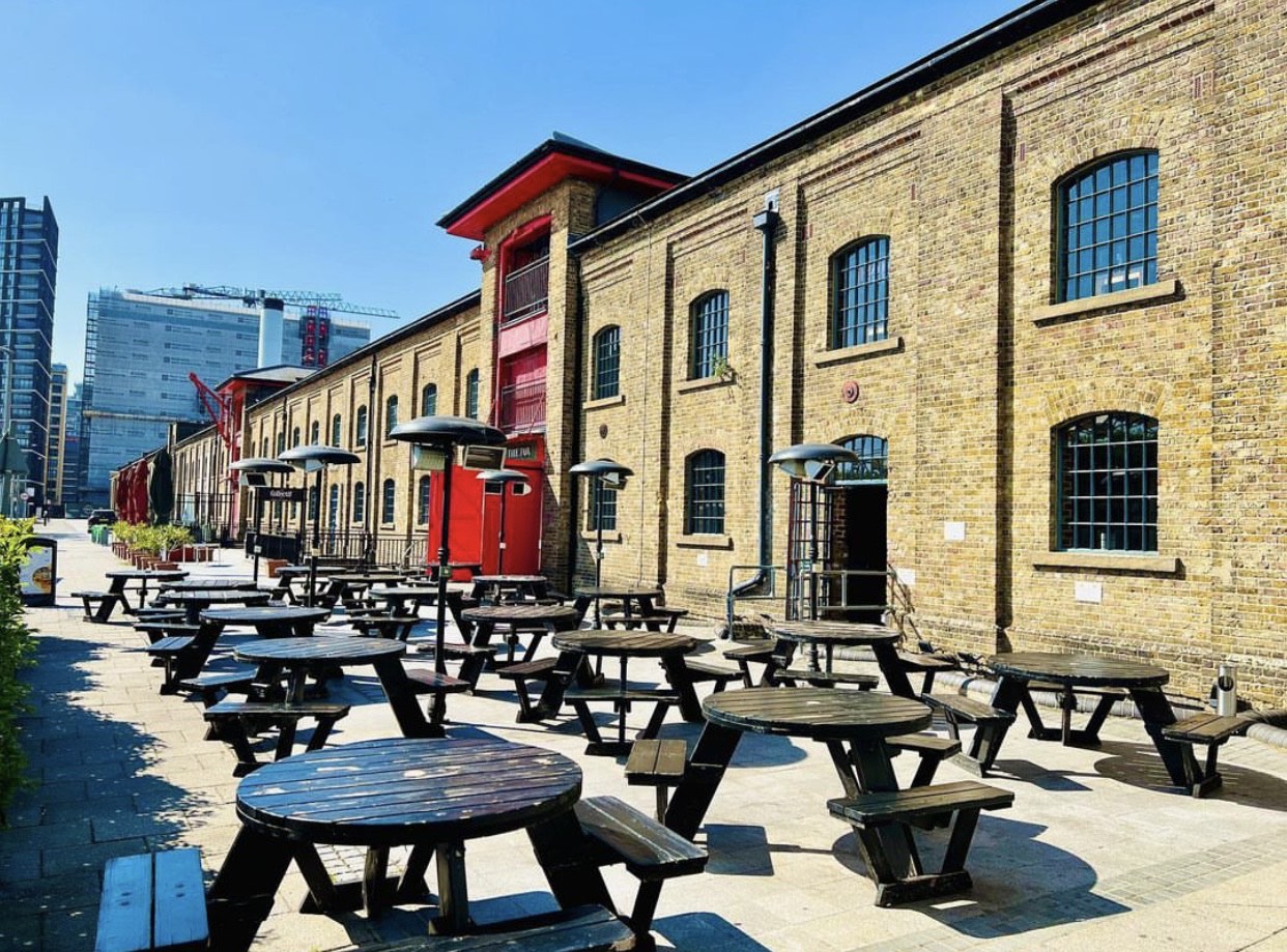 A snady coloured brick building with picnic tables outside on a sunny day with clear blue skies