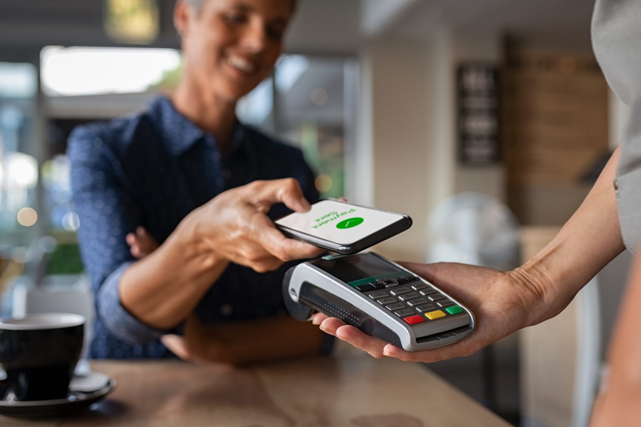 Mature woman paying bill through smartphone using NFC technology in a restaurant. Satisfied customer paying through mobile phone using contactless technology. Close up hands of mobile payment at a coffee shop.