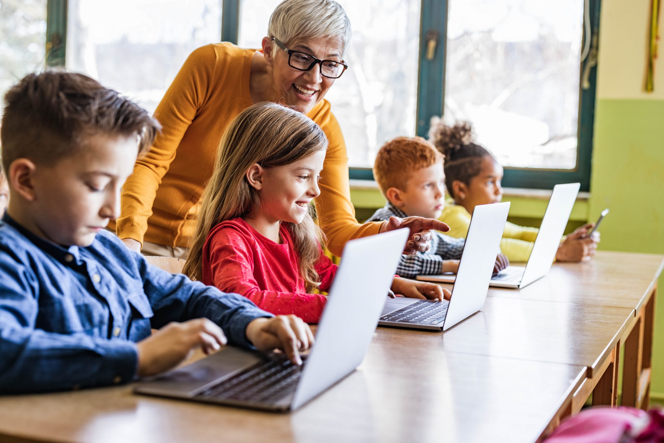 Happy mature teacher helping her students in using computers on a class at elementary school. Focus is on happy girl.