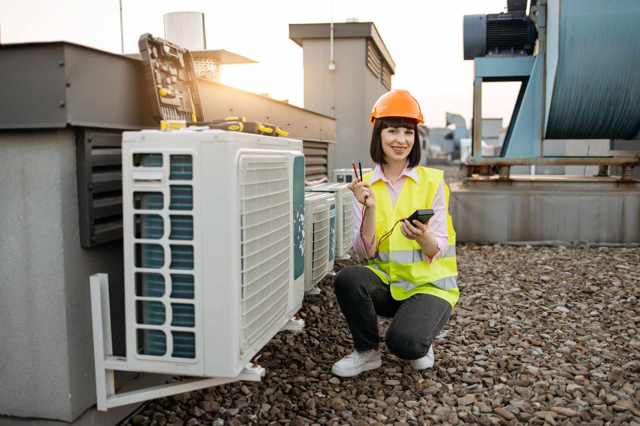 Female HVAC technician wearing a safety vest and hard hat, holding tools, and performing maintenance on an air conditioning unit on a rooftop.