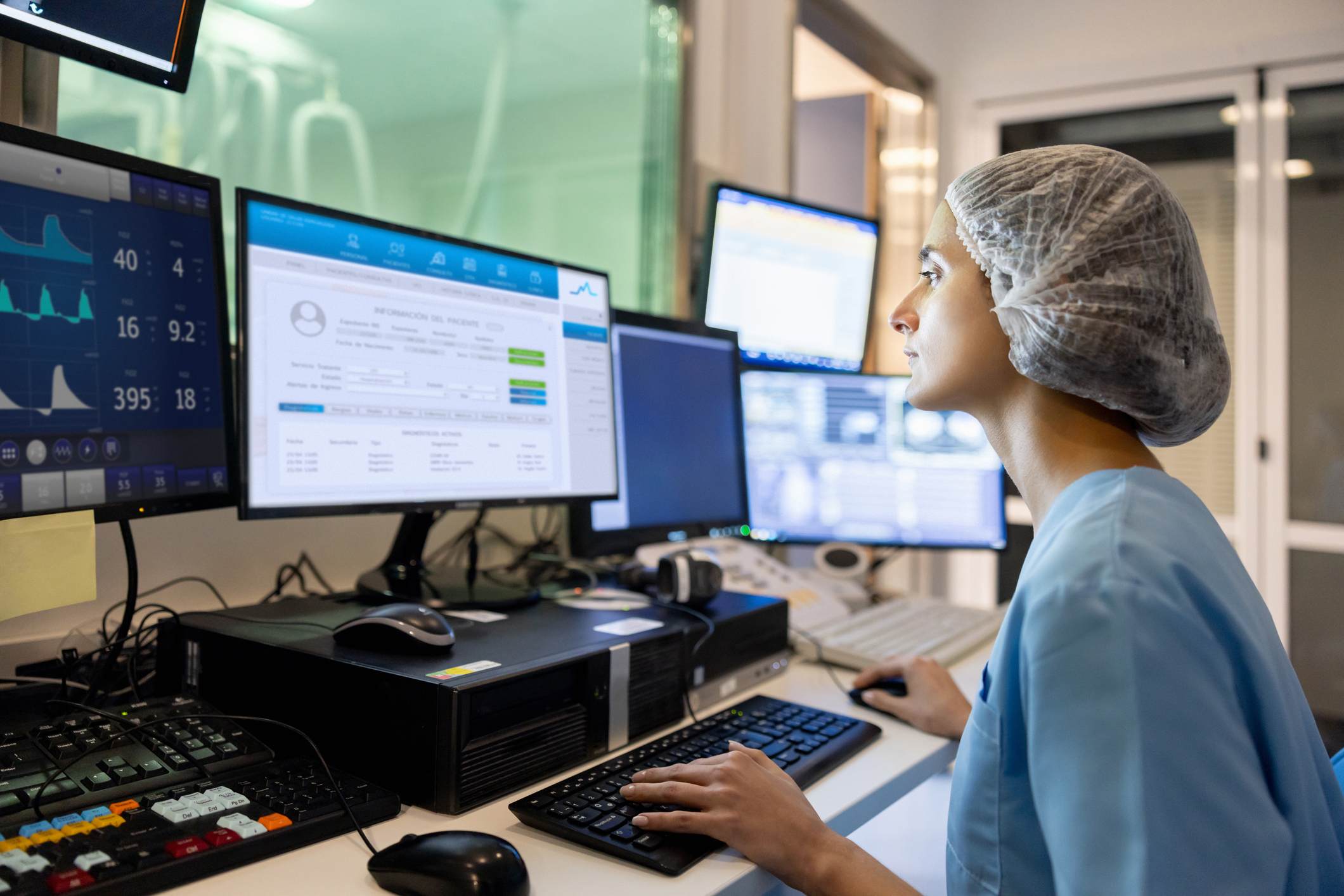 Female healthcare professional monitoring patient data on multiple computer screens in a hospital control room.